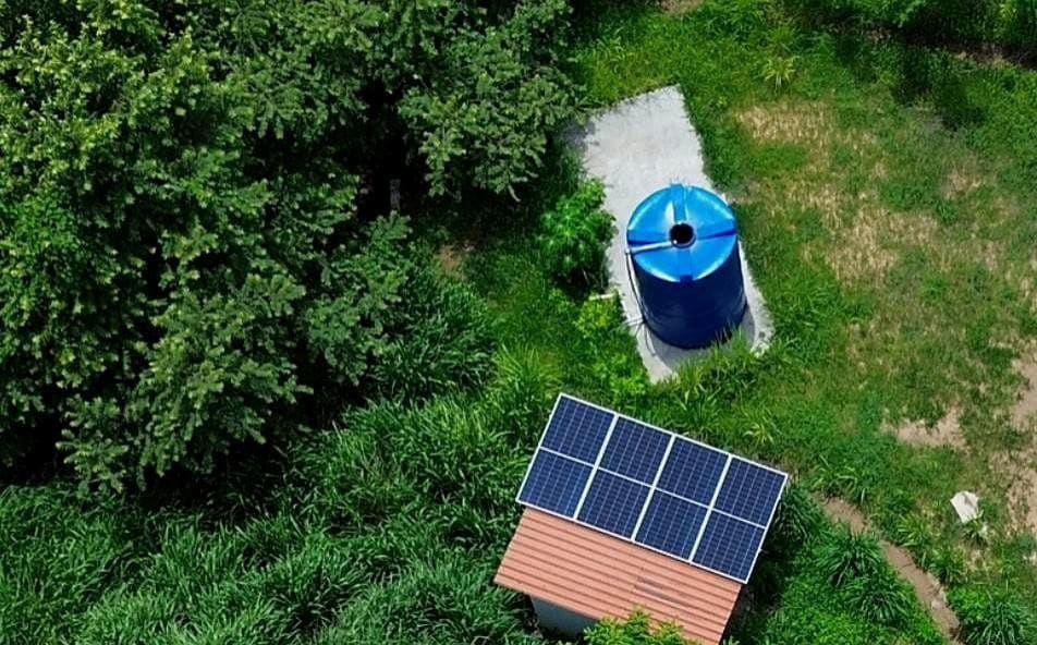 On site water pump housing with solar panels on its roof and a blue storage tank beside it